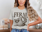White t-shirt with brown text "FERAL" and a raccoon wearing sunglasses.