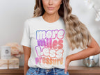 White t-shirt with "more miles less pressure" text in ombre gradient colors.