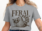 Grey t-shirt with brown "FERAL" text and raccoon wearing sunglasses.