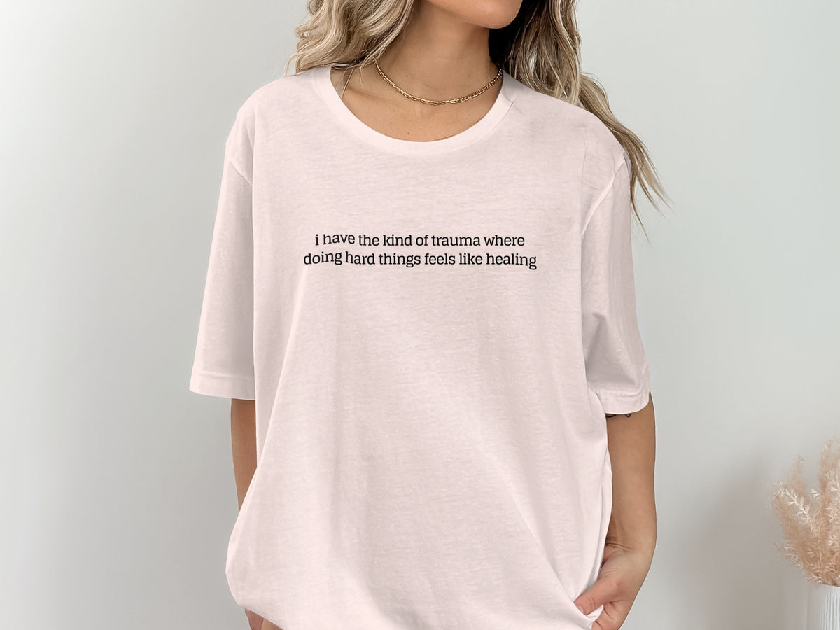 White t-shirt with text: "i have the kind of trauma where doing hard things feels like healing"