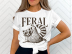 White t-shirt with "FERAL" and a raccoon wearing sunglasses.