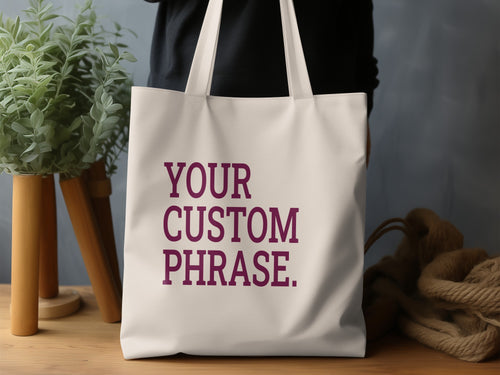 Cream tote bag with purple text "YOUR CUSTOM PHRASE."
