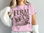 Pink t-shirt with "FERAL" text and a raccoon wearing sunglasses.