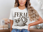 White t-shirt with "FERAL" text above a raccoon wearing sunglasses.