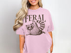 Pink t-shirt with "FERAL" text and a raccoon wearing sunglasses.